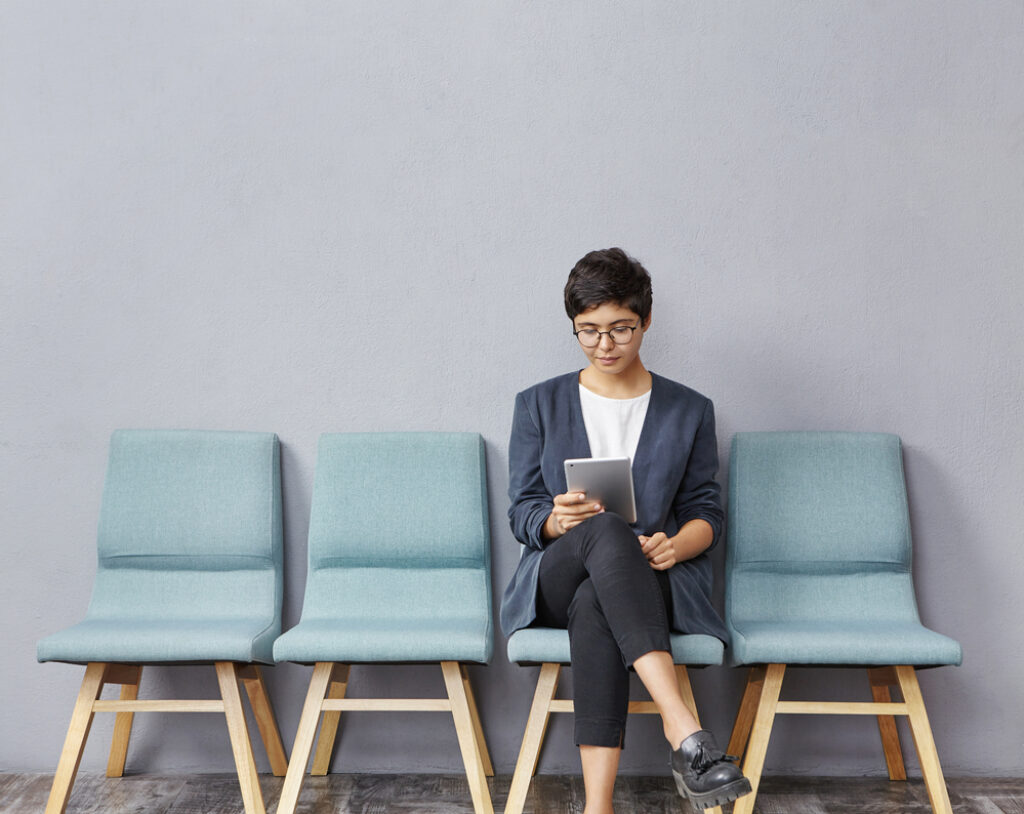 a lady sits on a seat in a row of seats in this hospital waiting room while  looking at her phone